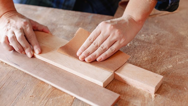 Person hand sanding a wooden plank along the direction of the grain using sandpaper.