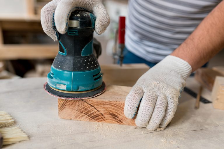 Close-up of a random orbital sander being used on a block of wood with gloves.