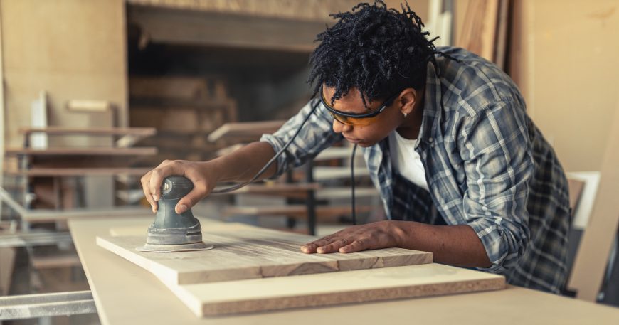 Woodworker using an orbital sander on a wooden board in a workshop.