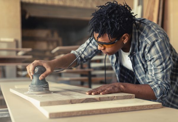 Woodworker using an orbital sander on a wooden board in a workshop.