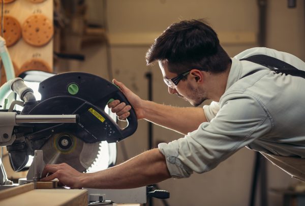 Young,Brunette,Man,In,Black,Overalls,By,Profession,Carpenter,Builder Woodworker using a circular saw to cut wood in a workshop
