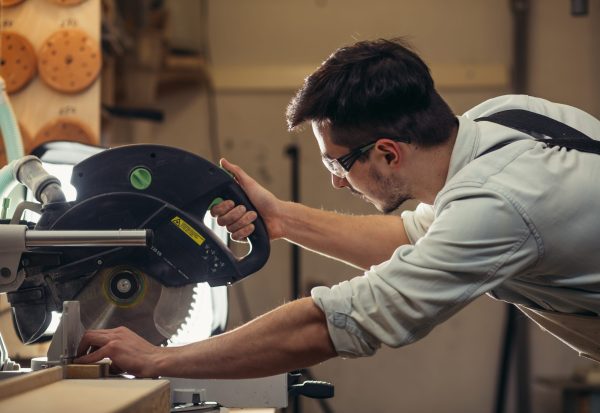 Young,Brunette,Man,In,Black,Overalls,By,Profession,Carpenter,Builder Woodworker using a circular saw to cut wood in a workshop