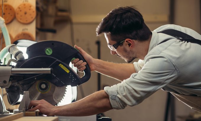Young,Brunette,Man,In,Black,Overalls,By,Profession,Carpenter,Builder Woodworker using a circular saw to cut wood in a workshop