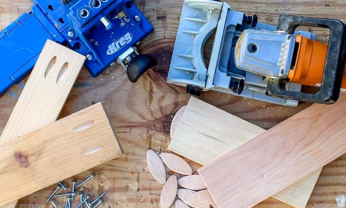 beginner-woodworking-joint-types-tools.png A top-down view of pocket hole jigs, a biscuit joiner, wood biscuits, and screws, illustrating common beginner woodworking joint types.