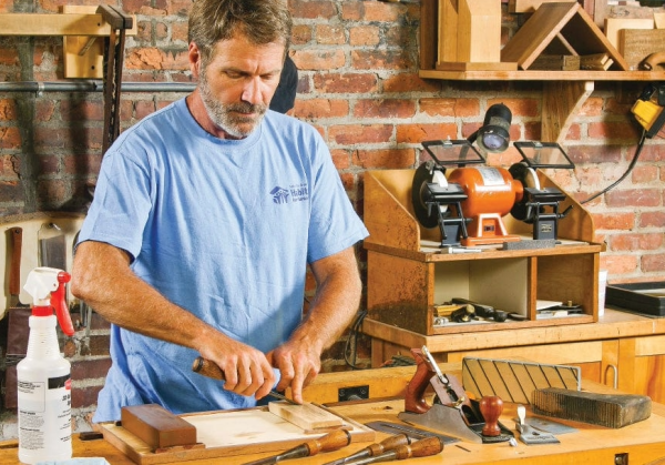 A woodworker using a sharpening stone at his workbench, demonstrating the setup for chisel and plane sharpening.