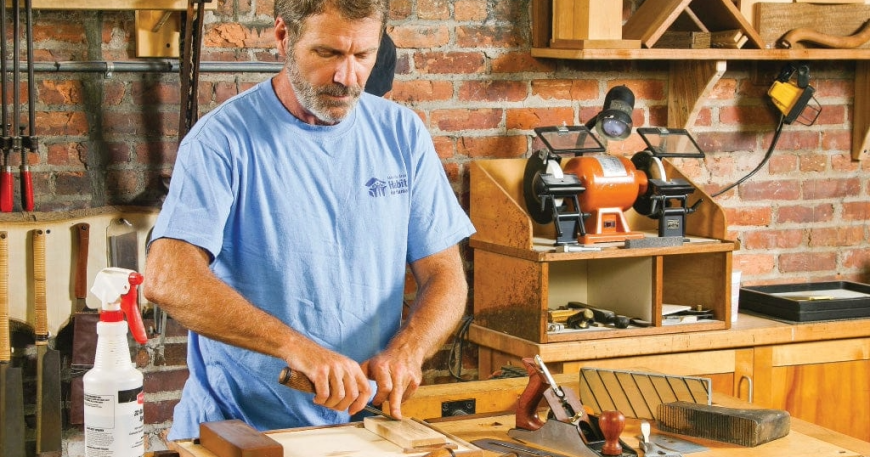 A woodworker using a sharpening stone at his workbench, demonstrating the setup for chisel and plane sharpening.