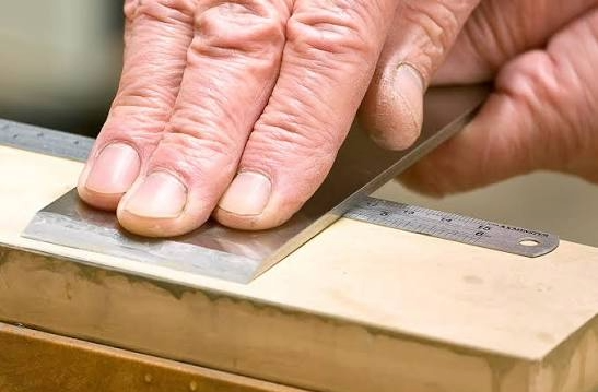 Close-up of a woodworker honing a hand plane blade on a waterstone using a thin steel ruler to flatten the back.