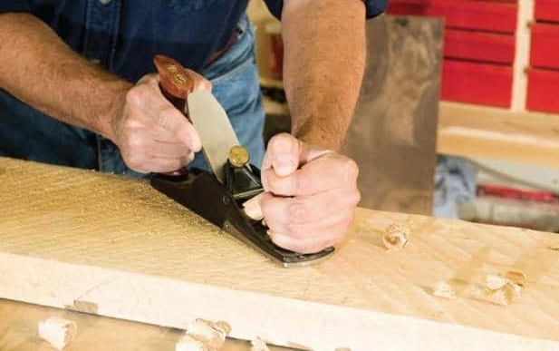 A woodworker using a hand plane on a wooden board, demonstrating the practical importance of planing wood with the grain.