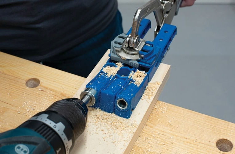 A close-up of a drill creating a pocket hole using a blue pocket hole joinery method guide clamp on a wooden board.