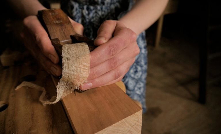 A wooden hand plane producing a thin, continuous wood shaving on a board, showcasing the result of proper sharpening.
